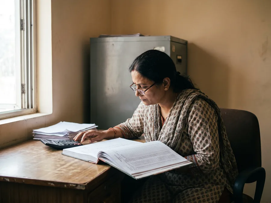 An accountant at her desk reading a printed ledger, a calculator and a stack of invoices beside her.